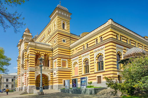 Pedestrian walks past the ornate facade of the Georgian National Opera and Ballet Theater in downtown Tbilisi, Georgia on a sunny day.