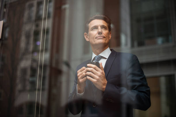 German businessman - looking through window Formally dressed 55-year-old businessman is standing in the bright office and looking through the window while holding a hot drink mug. georgijevic frankfurt stock pictures, royalty-free photos & images