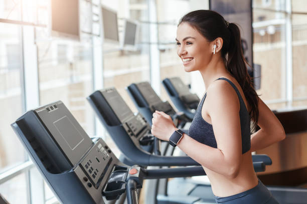 Get faster! Side view of young and cheerful woman in sportswear is running on a treadmill at gym and listening music. Cardio workout. stock photo