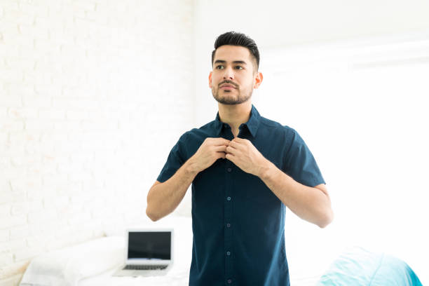 Getting Dressed In Bedroom Handsome young man buttoning his shirt in brightly lit room getting dressed stock pictures, royalty-free photos & images