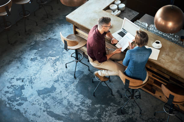 Getting work done at their local coffee shop High angle shot of two businesspeople working at the bar in a cafe coffee shop stock pictures, royalty-free photos & images