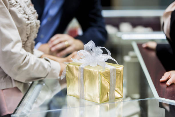 Gift box in jewelry store A gift box wrapped in gold wrapping paper, tied with a white bow, resting on the display case of a jewelry store. A saleswoman and customers, a couple, are cropped and out of focus in the background. store clerk selling jewelry stock pictures, royalty-free photos & images