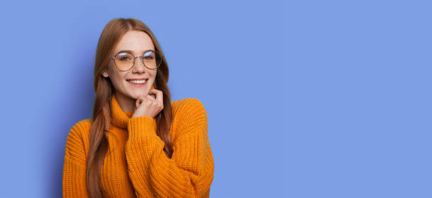 Ginger woman with red hair is smiling at camera with glasses on a blue studio wall with free space stock photo
