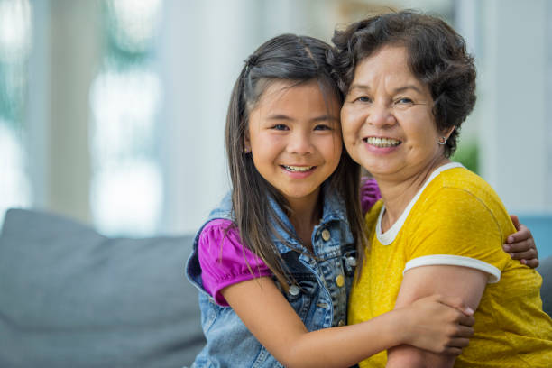 Girl And Grandmother An Asian family is indoors in a living room. They are wearing casual clothing. A girl and her grandmother are hugging while on a couch. filipino family stock pictures, royalty-free photos & images