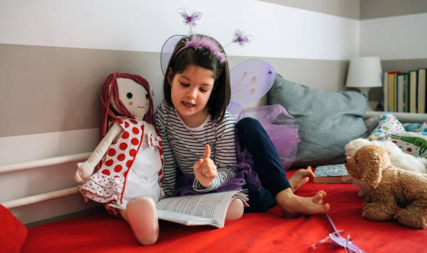 Girl disguised reading a book to her doll Little girl disguised as a butterfly sitting on the bed reading a book to her rag doll doll stock pictures, royalty-free photos & images
