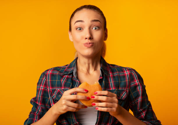 Girl Eating Burger Having Cheat Meal Standing On Yellow Background Slim Girl Eating Burger Having Cheat Meal Day Standing On Yellow Studio Background. Unhealthy Diet chewing stock pictures, royalty-free photos & images