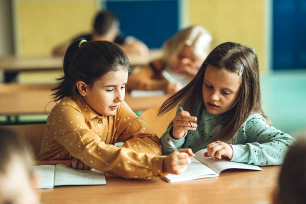 Girl explaining female friend while doing homework in classroom stock photo