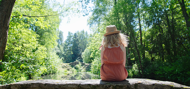 a little girl in an orange dress and hat is sitting on a stone bridge over a river in the forest, on a sunny summer day.  Panorama view