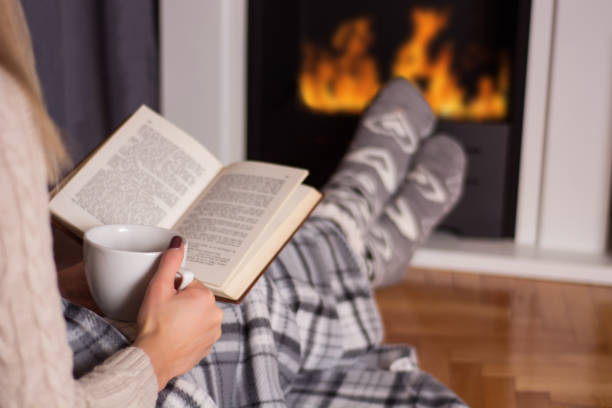 Girl in front of the fireplace reading book and warming feet on fire stock photo