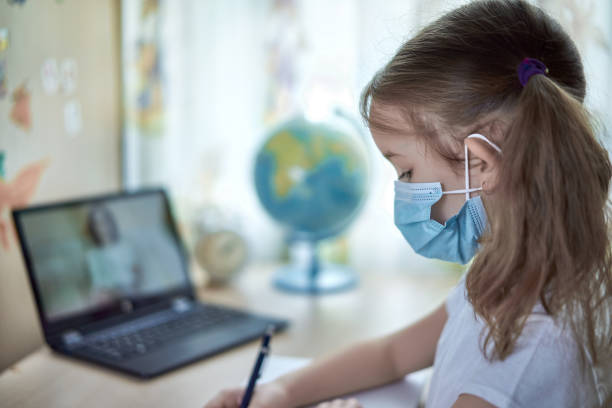 Girl in medical mask does homework at home stock photo