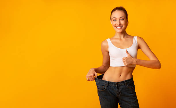 Girl In Oversize Jeans Gesturing Thumbs Up Smiling, Yellow Background Weight Loss. Girl In Oversize Jeans Gesturing Thumbs Up Smiling At Camera Standing On Yellow Studio Background. Copy Space thin stock pictures, royalty-free photos & images