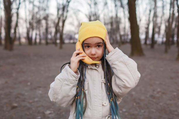 Girl in yellow hat talking on banana like cell phone in autumn forest, digital detox joke, lifestyle stock photo