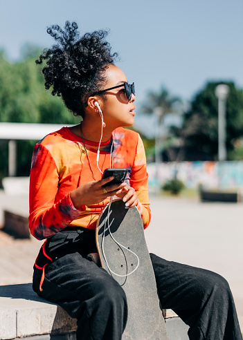 Young girl relaxing with her skateboard listening to music. Urban girl enjoying listening to music from her mobile phone while sitting at skatepark.