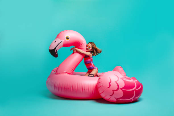 Girl on a giant inflatable flamingo imagining in the pool Girl in swimwear sitting on a giant inflatable pink flamingo. Small girl wearing swimsuit on a inflatable toy mattress over blue background. little girls in bathing suits stock pictures, royalty-free photos & images