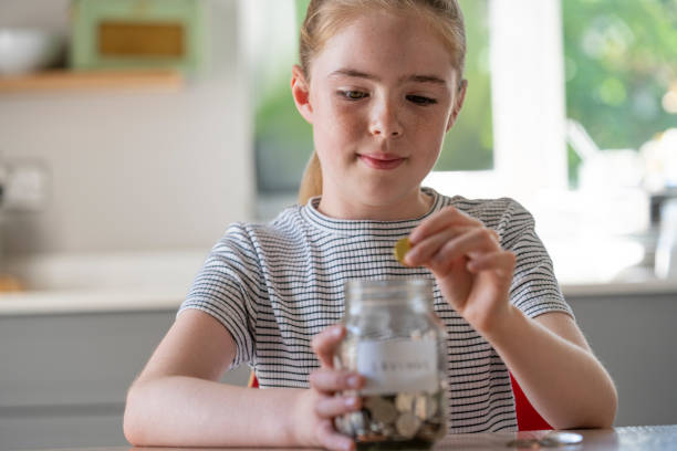 Girl Putting Coins Into Glass Jar Labelled Savings At Home Girl Putting Coins Into Glass Jar Labelled Savings At Home allowance stock pictures, royalty-free photos & images