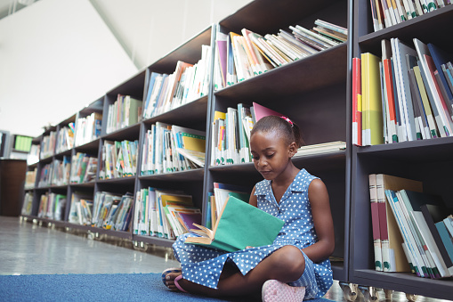 Girl reading book by shelf in library