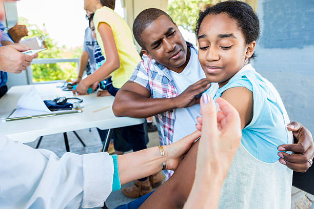 Girl receives flu shot at outdoor free clinic African American father comforts his preteen daughter while she receives a flu vaccine. The girl has a scared expression on her face. The nurse or doctor is giving her the injection in her arm. Patients are waiting in the background. free image of vaccinations stock pictures, royalty-free photos & images