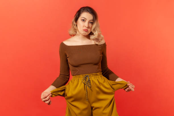 Girl showing empty pockets, no money Poor student, unemployment. Portrait of upset jobless woman in brown blouse turning out empty pockets, showing no money gesture, worried about debts. Indoor studio shot isolated on red background women pocket stock pictures, royalty-free photos & images