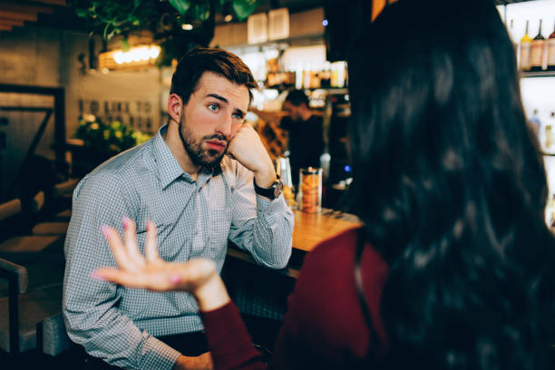 Girl sitting in front of young guy and talking to him. He looks bored. Man is not interested in conversation at all. stock photo