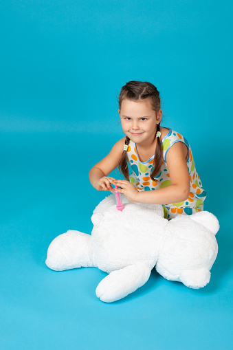 insidious, cunning girl sitting on the floor makes an injection or vaccination with a toy syringe in the ass of a white teddy bear isolated on a blue background.