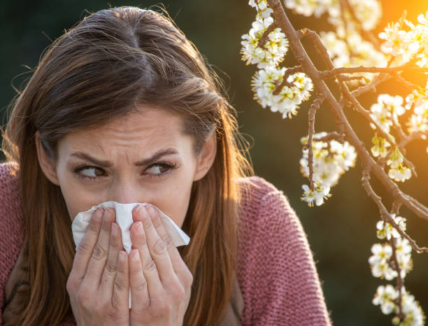 Girl sneezing in front of blooming tree in spring. Coronavirus or allergy Covid-19 or spring allergy. Afraid young woman looking at blooming tree and sneezing antihistamine stock pictures, royalty-free photos & images