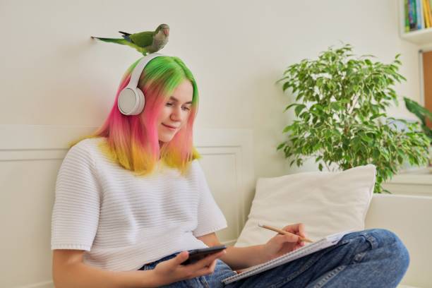 Girl student teenager sitting at home on bed with pet parrot on the head Girl student teenager sitting at home on bed with pet parrot on the head. Female student in headphones with school notebook and smartphone, e-learning pink hair stock pictures, royalty-free photos & images