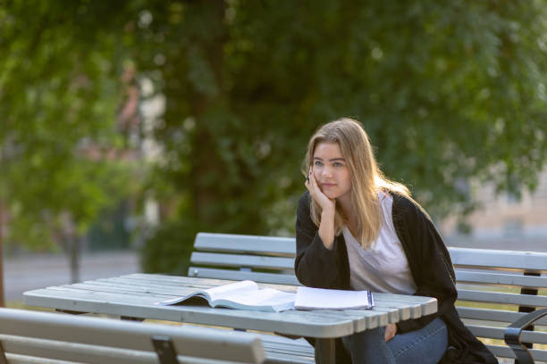 Girl studying in a park A girl is sitting on a bench in a park while studying. She is looking up and a textbook and a notepad are lying on the table in front of her. swedish girl stock pictures, royalty-free photos & images