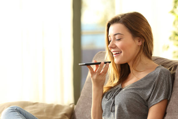 Girl using a smart phone voice recognition Girl using a smart phone voice recognition on line sitting on a sofa in the living room at home with a warm light and a window in the background speech recognition stock pictures, royalty-free photos & images