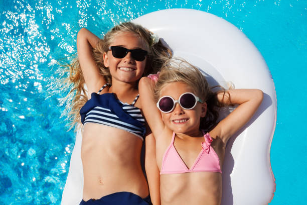 Girls friends enjoying summer in swimming pool Top view portrait of two age-diverse girls, happy friends in sunglasses and bikini, sunbathing on inflatable mattress in swimming pool little girls in bathing suits stock pictures, royalty-free photos & images