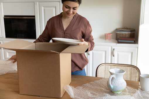 Unpacking with love. Glad young indian female shopper get new ordered kitchenware by mail open shockproof wrapping satisfied with no damage on dishes. Happy ethnic woman unbox postal parcel at kitchen
