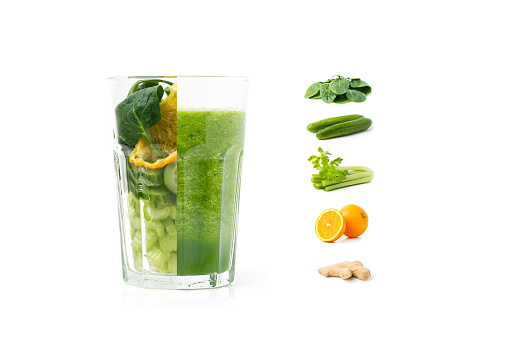 Detox cleanse drink concept. A glass filled with half smoothie and half colorful ingredients that go into the smoothie. Celery, cucumber, orange and spinach mix isolated on white.