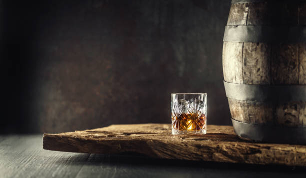 Glass of whisky or bourbon in ornamental glass next to a vinatge wooden barrel on a rustic wood and dark background stock photo