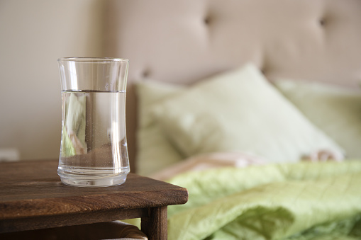 Transparent glass with water on a wooden nightstand in bedroom. Selective focus.