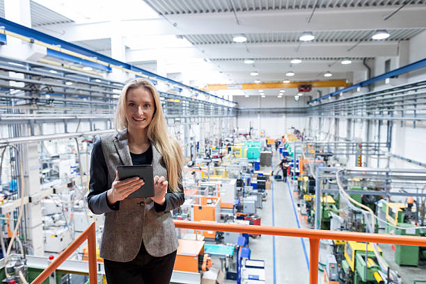 Global business communication in factory Horizontal color image of blond business female worker standing on balcony on top of large factory, holding digital tablet and examining the production online. Focus on attractive businesswoman sincerely smiling at camera, futuristic machines in background. Network Systems Administrator stock pictures, royalty-free photos & images