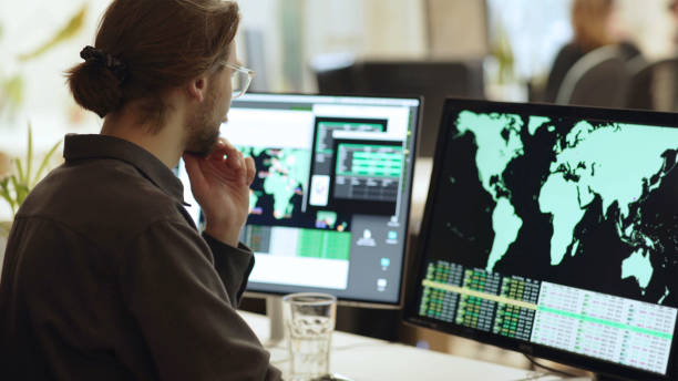 Global data office screens Stock image of a young bearded man, wearing glasses, surrounded by computer monitors in an office. In front of him there are screens showing maps of the world with associated data. person looking at map stock pictures, royalty-free photos & images