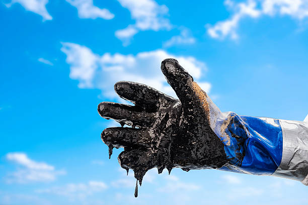Gloved hand of oil spill worker A close-up of a protective blue gloved hand of a clean up worker dripping with crude oil and sand during oil spill clean up against a partly cloudy sky tar stock pictures, royalty-free photos & images