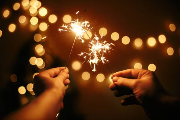 Glowing sparklers in hands on background of golden christmas tree lights, couple celebrating in dark festive room. Happy New Year. Space for text. Fireworks burning in hands. Happy Holidays stock photo
