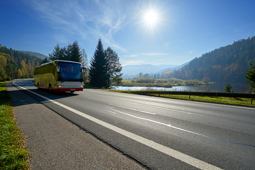 Glowing sun over the landscape with a bus traveling on the road around a lake and forested mountains in autumn