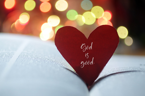Spiritual inspirational quote - God is love. With hand holding red valentines day card with heart shaped paper on an open page of bible book and colorful bokeh background.