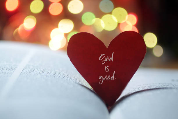 God is Good. Spiritual inspirational quote - God is love. With hand holding red valentines day card with heart shaped paper on an open page of bible book and colorful bokeh background. god stock pictures, royalty-free photos & images