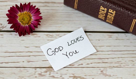 God Jesus Christ loves you handwritten message inspiring quote with closed Holy Bible Book, and flower on wooden background. Christian biblical concepts of love, faith, comfort, and peace. A close-up.