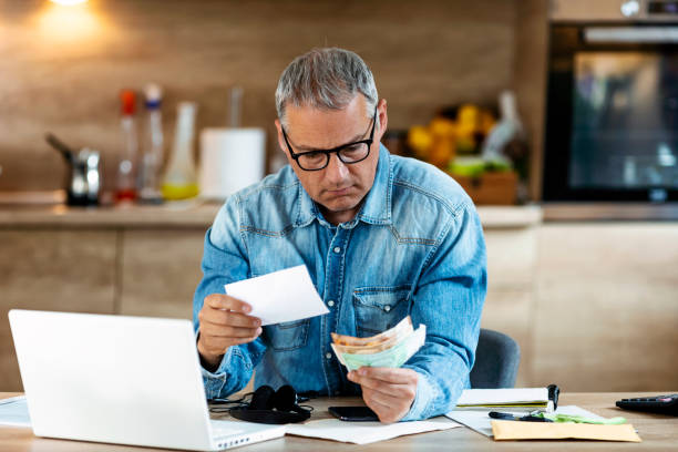 Going over the fine print may save your company Confused frustrated mature man holding mail letter and money in the kitchen, reading shocking unexpected news nonsense in paper document, mad about high bill tax invoice, debt notification, bad financial report, money problem. bankruptcy stock pictures, royalty-free photos & images