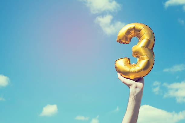 Gold number 3 balloon A gold foil number 3 balloon is held high in the air by caucasian male hand.  The image has been taken outdoors on a bright sunny day, the sky is blue with some clouds. A vintage style effects has been added to the image. 2 3 years stock pictures, royalty-free photos & images