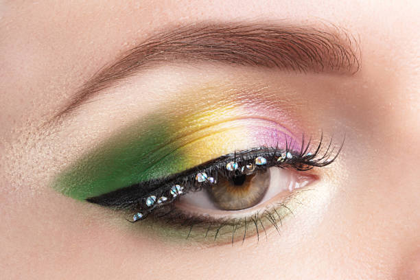 Gold, purple and green Mardi gras makeup. Woman closed eye with bright eyeshadows and rhinestones on false eyelashes closeup Gold, purple and green Mardi gras makeup. Woman closed eye with bright eyeshadows and rhinestones on false eyelashes closeup. mardi gras women stock pictures, royalty-free photos & images