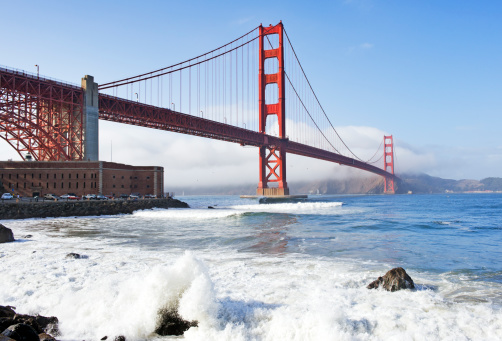 This view of the Golden Gate Bridge takes in the historic Ft Point as well as the sun shining thru this fantastic bridge with the North end with fog and the waves crashing against the rocks and shoreline.