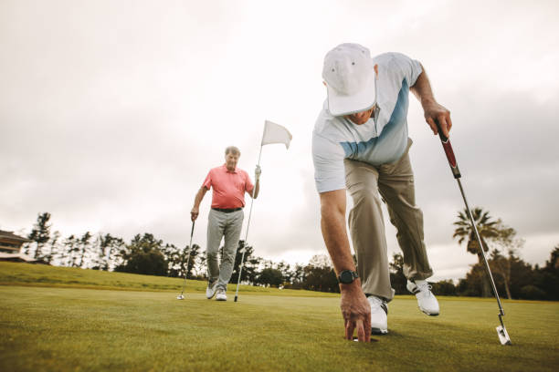 Golfer picking golf ball out of hole Senior golf player picking golf ball out of hole with second player in the background holding the flag. Senior golf players playing a game on the golf course. bending stock pictures, royalty-free photos & images