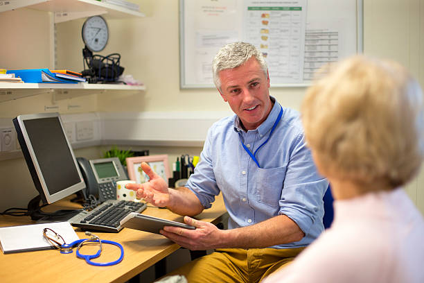 Good Bill of Health A male doctor sits face to face with an elderly woman. They are in the doctors office . The focus is on the doctor who sits at his desk holding a digital tablet and smiling at the woman. The woman face cannot be seen. general practitioner stock pictures, royalty-free photos & images