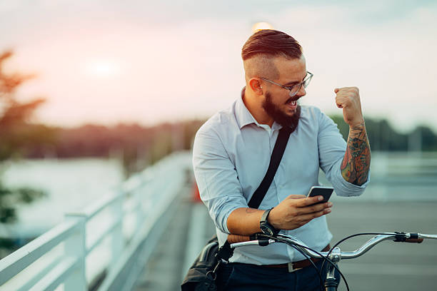 Good Business News! Young Businessman pushing his city bicycle and using his smart phone. He has tattoos and hipster haircut. He just got positive reports from stock market. Using app on his smart phone. He is celebrating his success with fist up. good news stock pictures, royalty-free photos & images