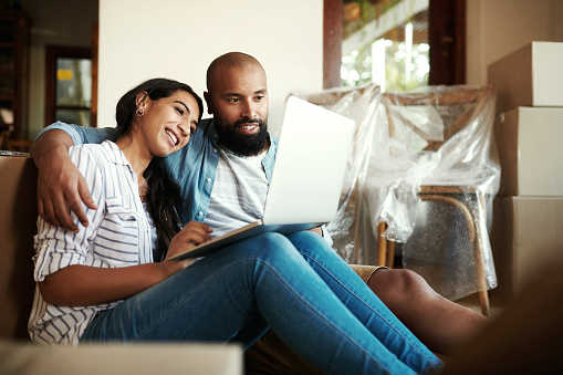 Shot of a young smiling couple sitting on the floor surrounded by moving boxes while they use a laptop