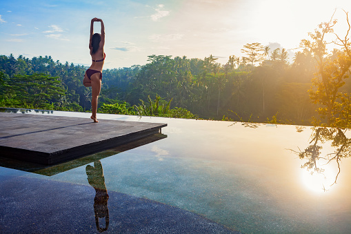 Good morning with yoga meditating on sunrise background. Active woman in bikini practicing on villa poolside to keep fit and health. Woman fitness training, sport activity on summer family holiday.